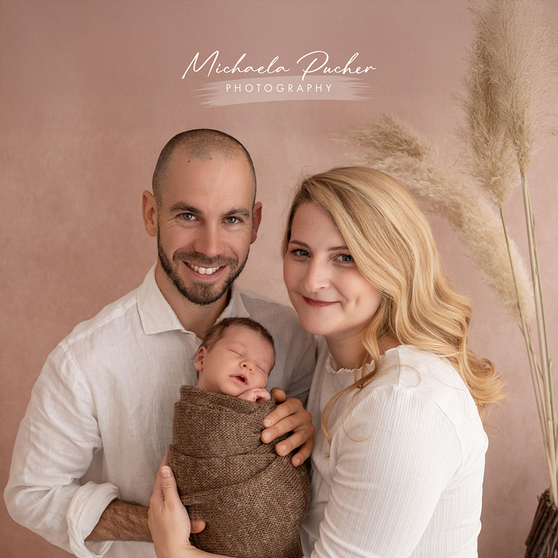 Ein Paar hält sein schlafendes Neugeborenes gemeinsam im Arm; alle tragen helle Kleidung und stehen vor einem warmen, rosigen Hintergrund mit Pampasgras.